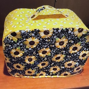 May include: A quilted bread maker cover with a black background and a pattern of yellow sunflowers and bees. The cover has a yellow handle and a yellow border.