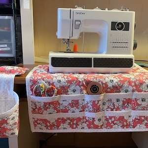 May include: A white sewing machine with a pink and white floral fabric sewing machine table organizer with multiple pockets. The organizer has a white trim with black polka dots. The sewing machine has the text "Strong & Tough" on the front.