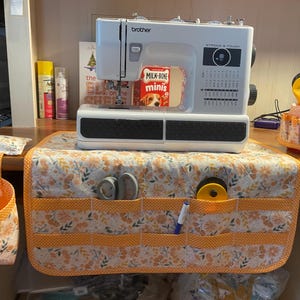 May include: A white sewing machine with a floral fabric organizer with multiple pockets. The organizer has a pocket for scissors, a pocket for a spool of thread, and a pocket for a pen. The sewing machine is on a wooden table with a variety of items on it, including a box of Milk-Bone dog treats.