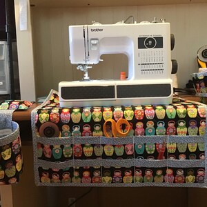 May include: A white Brother sewing machine sits atop a sewing table organizer with pockets. The organizer and matching accessories feature a black background with colorful Matryoshka doll designs. Scissors and rotary cutter are visible.