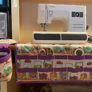 May include: A white Brother sewing machine sits atop a sewing table with a colorful fabric organizer. The organizer features pockets and a print of jars filled with sewing notions. A purple polka-dot fabric basket hangs on the left.