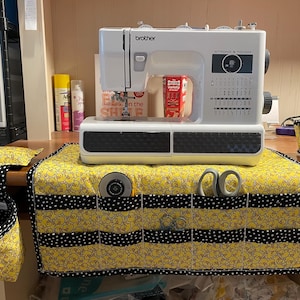 May include: A white Brother sewing machine sits atop a yellow and black duck-themed sewing machine mat with pockets. A matching hanging basket and pincushion are on the left. Scissors and a rotary cutter are in the pockets.