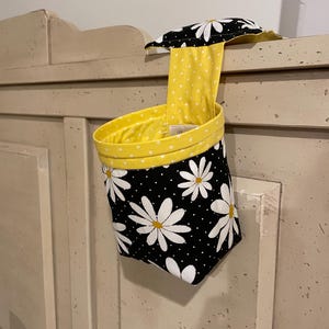 Puede incluir: Un contenedor de almacenamiento de tela negro y blanco con un forro de lunares amarillos y un estampado de margaritas. El contenedor está colgado en una puerta de armario blanca.