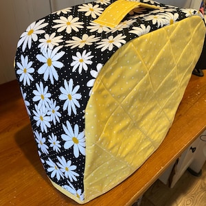 May include: A yellow and black quilted fabric cover with a daisy print and a handle. The cover is designed to fit a kitchen appliance.