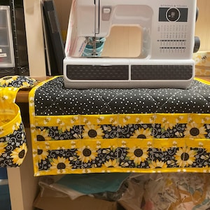 May include: A white sewing machine sits on a black and white polka dot quilted mat with a sunflower and bee print border. The mat features multiple storage pockets. A matching hanging fabric basket and a small pincushion are attached to the side.