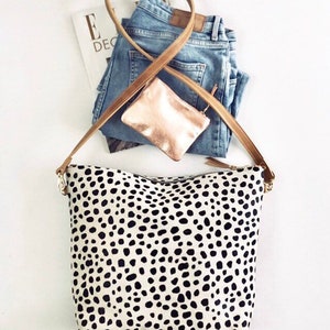 May include: A cream-colored tote bag with a black dalmatian spot pattern, a brown leather strap, and gold-colored hardware. The bag is paired with blue jeans, a small rose gold pouch, and a magazine.