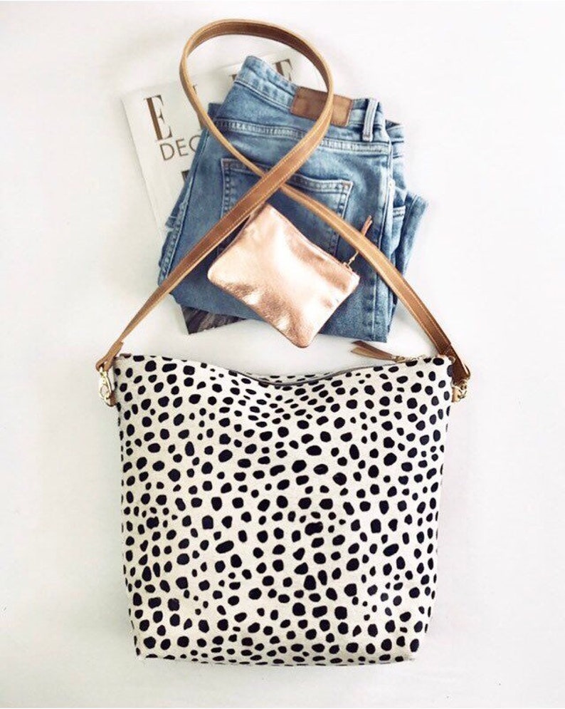 May include: A cream-colored tote bag with a black dalmatian spot pattern, a brown leather strap, and gold-colored hardware. The bag is paired with blue jeans, a small rose gold pouch, and a magazine.