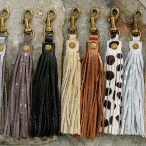 May include: Ten leather keychains with tassels in various colours and patterns, including leopard print, black, grey, white, gold, and brown. Each keychain has a gold metal clasp.