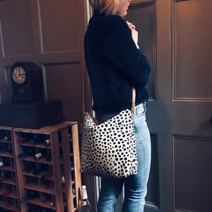 May include: A black and white spotted crossbody bag with a light brown strap. The bag is worn over a dark blue sweater and blue jeans. The bag's pattern is reminiscent of a Dalmatian dog.
