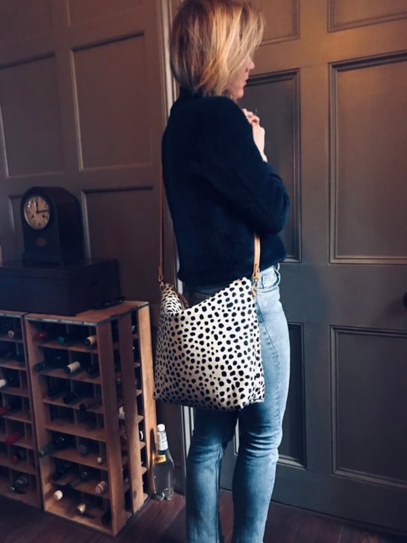 May include: A black and white spotted crossbody bag with a light brown strap. The bag is worn over a dark blue sweater and blue jeans. The bag's pattern is reminiscent of a Dalmatian dog.