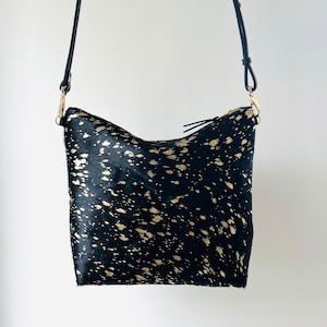 May include: Black and gold crossbody bag with a leather strap. The bag features a black background with gold splatter-like accents. The bag has a zipper closure and gold-colored hardware.