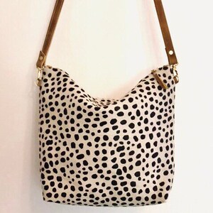 May include: A cream-colored shoulder bag with a black spotted pattern. The bag has brown leather straps and gold-colored metal hardware. The bag is a medium size and appears to be made of fabric.