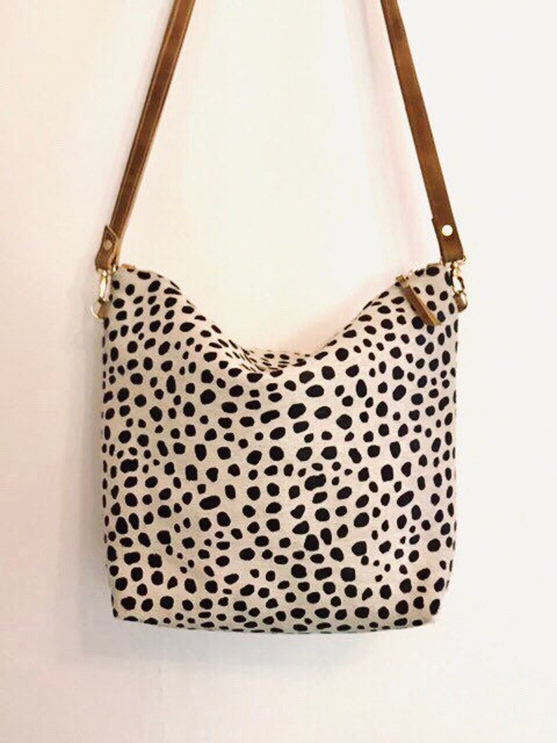 May include: A cream-colored shoulder bag with a black spotted pattern. The bag has brown leather straps and gold-colored metal hardware. The bag is a medium size and appears to be made of fabric.