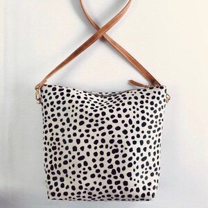 May include: A cream-colored tote bag with a black spotted pattern. The bag has a brown leather strap and gold-colored hardware. The bag is designed to be worn over the shoulder.