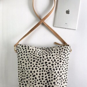 May include: A cream-colored handbag with a black spotted pattern. The bag has a tan leather strap and gold-colored zipper hardware. An iPad is visible in the background.