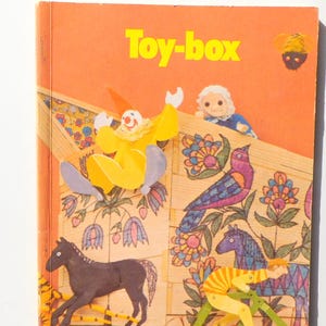 May include: Vintage children's book titled "Toy-box" with a bright orange cover. The cover features a colorful illustration of a clown, a bird, a horse, and a person riding a bicycle. The book has a retro design.