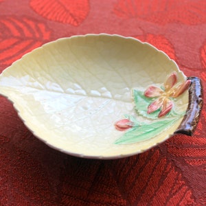 Puede incluir: Un plato de cerámica con forma de hoja, de color amarillo pálido, con una superficie texturizada. El plato está adornado con un diseño floral en tonos rosa, verde y marrón. El plato está colocado sobre una superficie roja estampada.