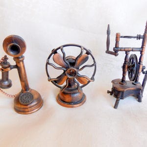 Op de afbeelding: Drie miniatuur objecten in antieke stijl: een draaitelefoon, een ventilator en een spinnewiel. De objecten zijn gemaakt van koperkleurig metaal en zijn geplaatst op een wit oppervlak. De telefoon heeft een ketting en de ventilator heeft bladen.