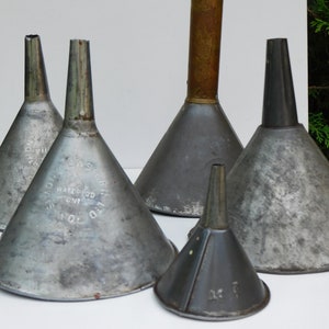 Puede incluir: Una colección de embudos de metal vintage de varios tamaños. Los embudos son de metal color plata, algunos muestran signos de edad y desgaste. Un embudo tiene una parte superior de latón. Los embudos están dispuestos sobre una superficie blanca.