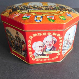 Puede incluir: Una lata octogonal roja con ilustraciones históricas. La tapa muestra una escena de reunión con texto. Los lados presentan retratos y escenas históricas, con varias banderas y diseños florales.