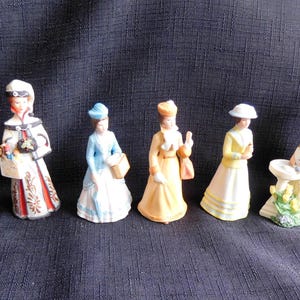 Può includere: Una collezione di sei statuine in porcellana raffiguranti donne in abiti d'epoca. Le statuine presentano una varietà di abiti e cappelli, con dettagli come cestini, fiori e accessori. La tavolozza dei colori comprende sfumature di grigio, bianco, blu, giallo e arancione.