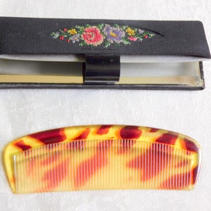 May include: A black compact with a floral embroidery design and a mirror inside. A tortoiseshell comb with a red and yellow pattern.