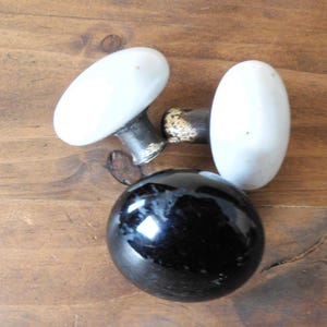 May include: A collection of vintage cabinet knobs and pulls. The image features two white oval knobs, a black round knob, and a knob with a dark metal base. The knobs are arranged on a wooden surface.