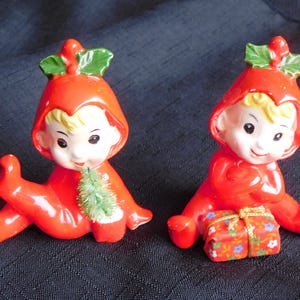 Peut inclure: Deux figurines en céramique d'elfes assis, vêtus de tenues à capuche rouges. L'un tient un petit sapin de Noël vert, l'autre un cadeau emballé. Les elfes ont les cheveux blonds et sont décorés de feuilles vertes et de baies rouges.