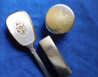 May include: An antique vanity set featuring a hand mirror, a compact, and a rectangular case. The mirror has a floral design and gold trim. The compact and case are gold-toned. The set is displayed on a blue surface.