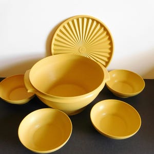 Puede incluir: Un juego de utensilios de cocina de plástico color amarillo mostaza que consta de un tazón grande para mezclar con dos asas laterales, cuatro tazones más pequeños a juego y una tapa circular con un patrón de líneas radiantes.