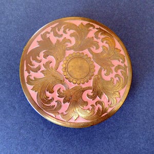 Op de afbeelding: Een ronde, roze compact met een gouden, sierlijk ontwerp van draaiende bladeren en een centrale cirkel.