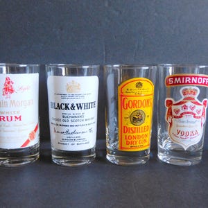 May include: Four clear glass shot glasses with different liquor brands printed on them. The glasses are arranged in a row from left to right: Captain Morgan White Rum, Black & White Scotch Whisky, Gordon's London Dry Gin, and Smirnoff Vodka.