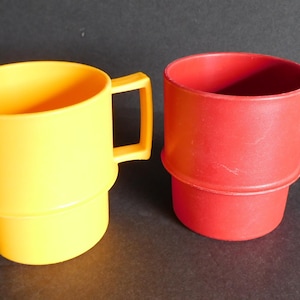Puede incluir: Dos tazas de plástico, una amarilla y otra roja, se muestran juntas. Cada taza tiene un asa y una base escalonada. Las tazas están hechas de un material plástico opaco y sólido. El fondo es de color oscuro y neutro.