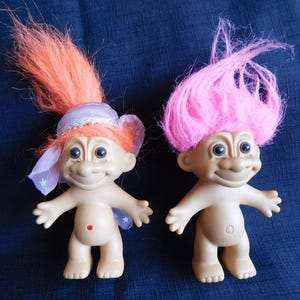 May include: Two vintage troll dolls with smiling faces and brightly colored hair. One doll has orange hair and a purple hat with a veil, the other has pink hair. Both dolls have a beige skin tone and are standing.
