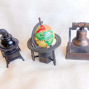 May include: Three miniature metal sculptures: a black stove, a globe on a stand, and a bronze Liberty Bell. The globe features a colorful map. The stove has a curved chimney. The bell is framed by a rectangular structure.