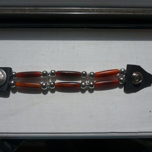 Puede incluir: Un brazalete de cuero marr&oacute;n con cuentas plateadas y cuentas cil&iacute;ndricas rojas y marrones. El brazalete tiene una moneda de plata con el perfil de un rostro masculino.
