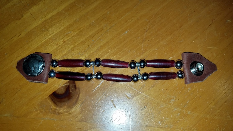 Puede incluir: Un brazalete de cuero marr&oacute;n con cuentas plateadas y cuentas de madera roja. El brazalete tiene un cierre en forma de moneda en un extremo y un cierre a presi&oacute;n en el otro extremo.