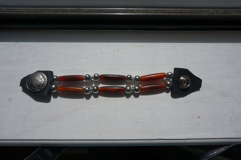 Puede incluir: Un brazalete de cuero marr&oacute;n con cuentas plateadas y cuentas cil&iacute;ndricas rojas y marrones. El brazalete tiene una moneda de plata con el perfil de un rostro masculino.