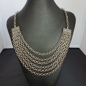 May include: Silver-toned necklace with multiple strands of chain. The necklace is displayed on a black mannequin. The chains are connected at the top with decorative rectangular pieces. The necklace has a layered design.
