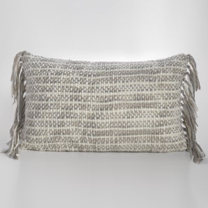 May include: A long rectangular throw pillow with a woven texture in shades of white and gray. The pillow has fringe on both ends.