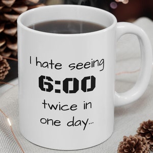 May include: White ceramic coffee mug with black text that reads "I hate seeing 6:00 twice in one day..." The mug is sitting on a white surface with a string of lights and pine cones.