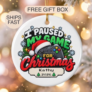May include: A round white ceramic ornament featuring a black game controller wearing a red and white Santa hat. The text reads I PAUSED MY GAME FOR Christmas, with the name Kathy and the year 2026 printed below in a pixelated font style.