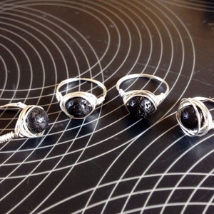 May include: Four handmade rings with a silver wire design. Each ring features a dark, round, porous stone. The rings are displayed on a black surface with a white grid pattern. The rings are a unique jewelry item.