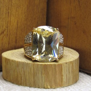 May include: A gold ring with a large, clear, rectangular-shaped gemstone. The gemstone has a slight yellow hue and is surrounded by smaller, clear gemstones.