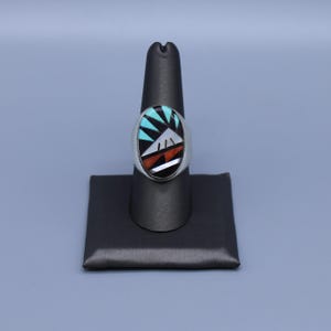 May include: A silver ring featuring an oval inlay design with geometric patterns. The inlay consists of turquoise, black, white, and orange stone segments arranged in a sunburst and landscape motif, set within a polished silver band frame.