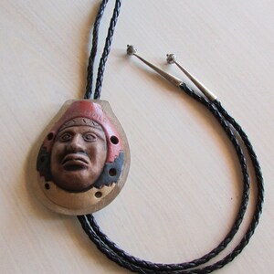 May include: A bolo tie with a ceramic pendant featuring a stylized face with the word "CHILE" inscribed on the top. The pendant is brown, red, and black with a white background. The bolo tie cord is black.