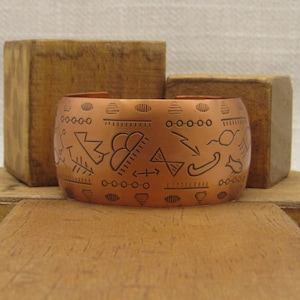 May include: A copper bracelet with an etched design of geometric shapes and symbols. The bracelet is a wide band with a rounded edge.
