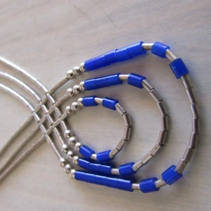 May include: A layered necklace featuring silver-toned metal chains and bright blue cylindrical beads. The necklace has a three-strand design with a cascading effect. The beads are evenly spaced along the strands, creating a modern and stylish look.