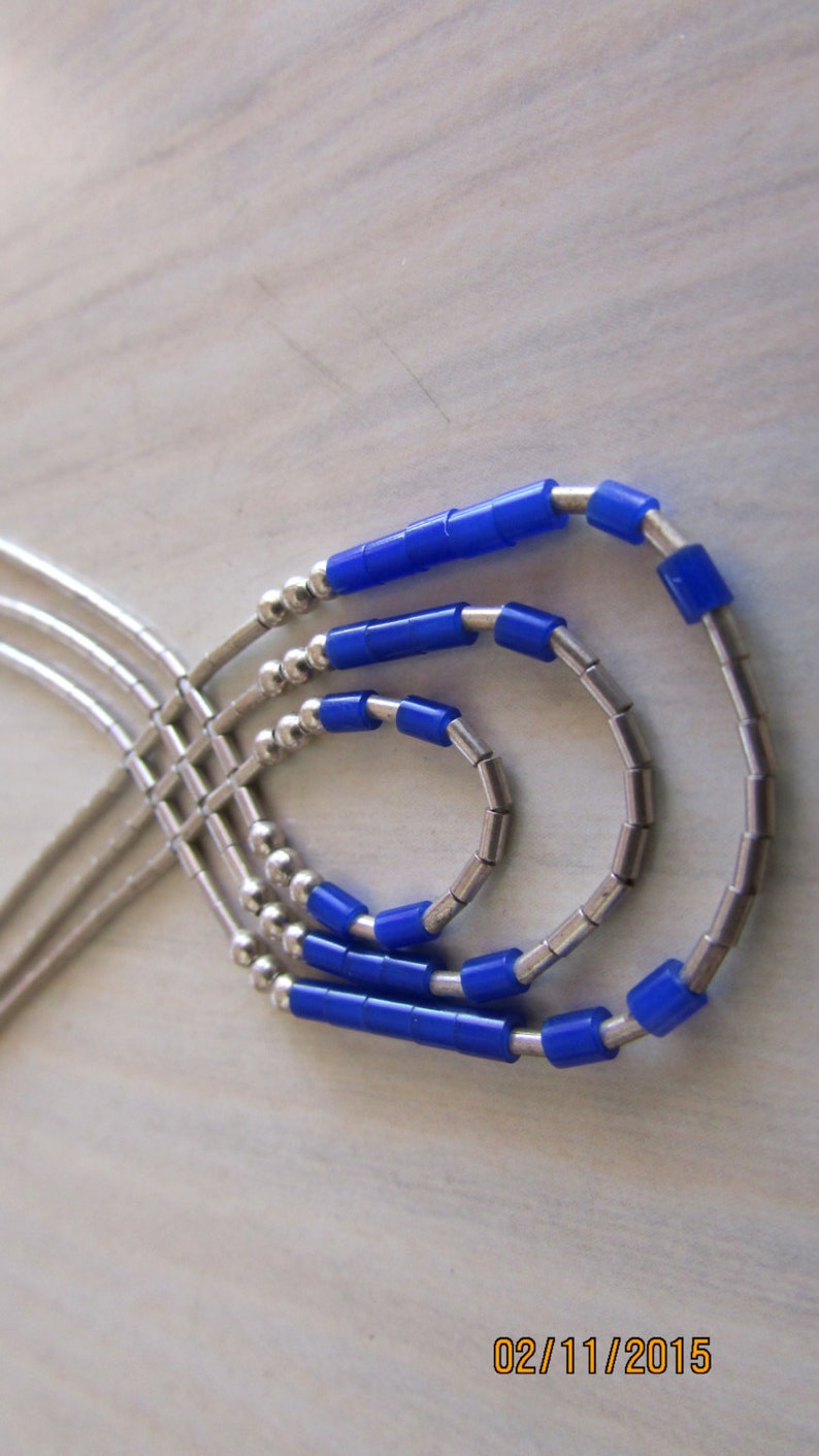 May include: A layered necklace featuring silver-toned metal chains and bright blue cylindrical beads. The necklace has a three-strand design with a cascading effect. The beads are evenly spaced along the strands, creating a modern and stylish look.