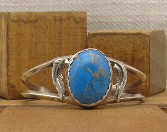 May include: A silver bracelet with a blue turquoise stone set in a silver oval bezel. The bracelet has two silver feather designs on either side of the stone.